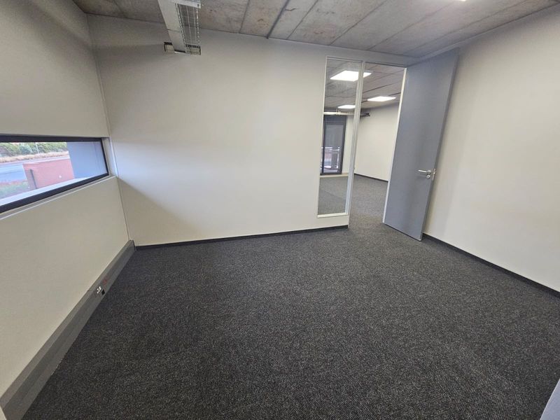 A clean, empty office space featuring exposed concrete ceilings, grey carpeting, and a glass partition wall, offering a modern and professional work environment.