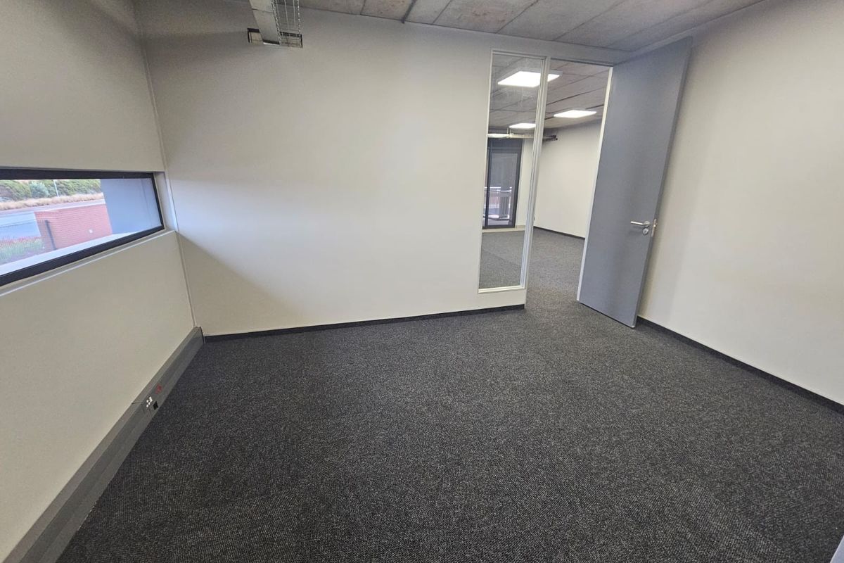 A clean, empty office space featuring exposed concrete ceilings, grey carpeting, and a glass partition wall, offering a modern and professional work environment.