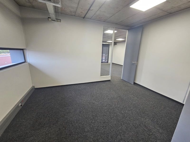 A clean, empty office space featuring an exposed concrete ceiling, recessed lighting, grey carpeting, and a glass partition leading to an adjacent room.