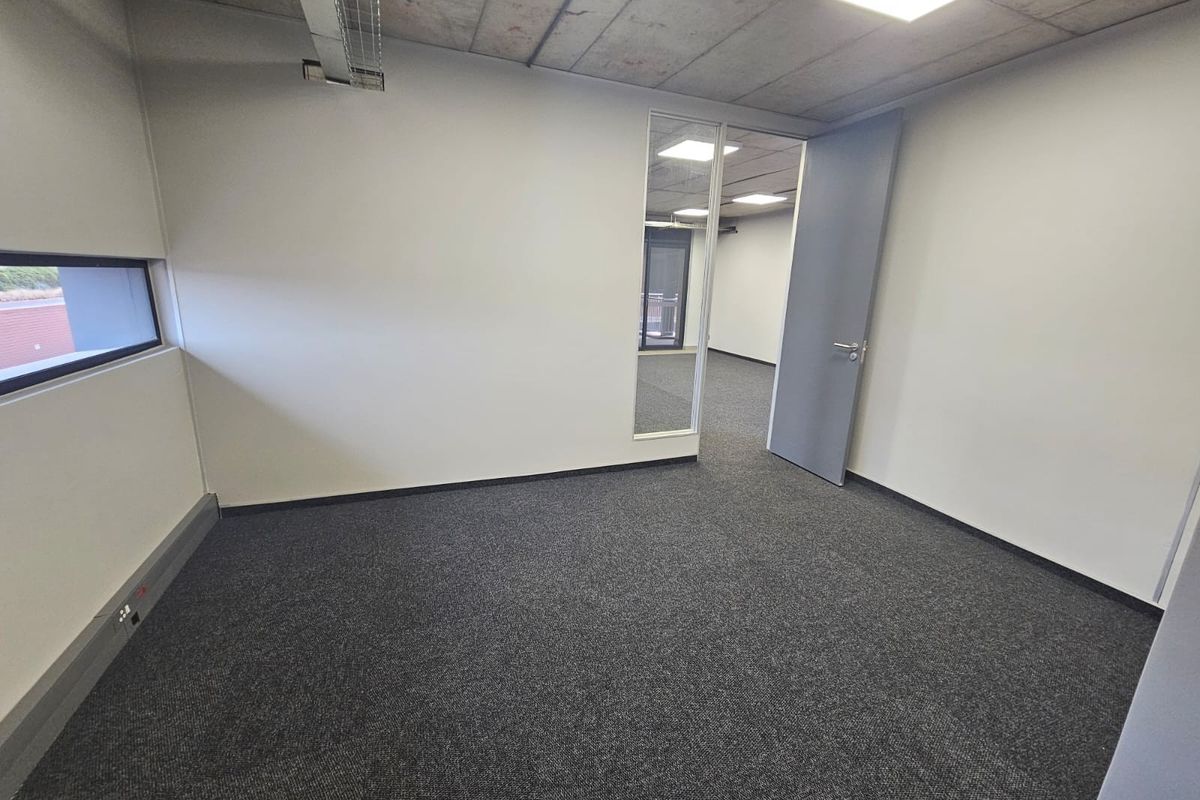 A clean, empty office space featuring an exposed concrete ceiling, recessed lighting, grey carpeting, and a glass partition leading to an adjacent room.