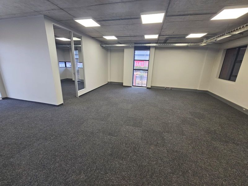A modern, open-plan office space featuring exposed concrete ceilings, recessed LED lighting, carpeted flooring, and a glass-partitioned private office area.
