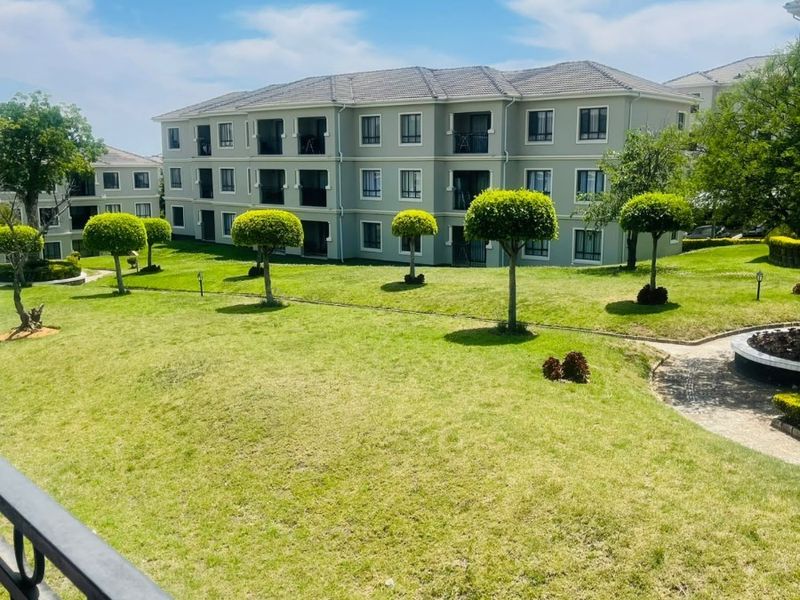 A view of a well-maintained apartment complex exterior featuring landscaped lawns, manicured trees, and a paved walkway, viewed from an elevated balcony.