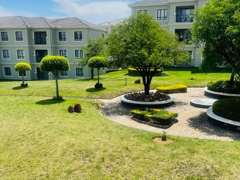 A well-maintained communal garden area featuring manicured lawns, paved walkways, and established trees, set against the backdrop of modern apartment buildings.