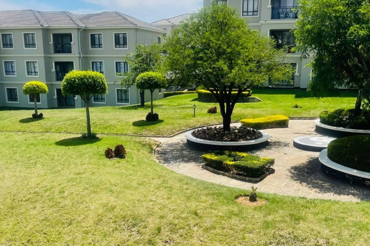 A well-maintained communal garden area featuring manicured lawns, paved walkways, and established trees, set against the backdrop of modern apartment buildings.