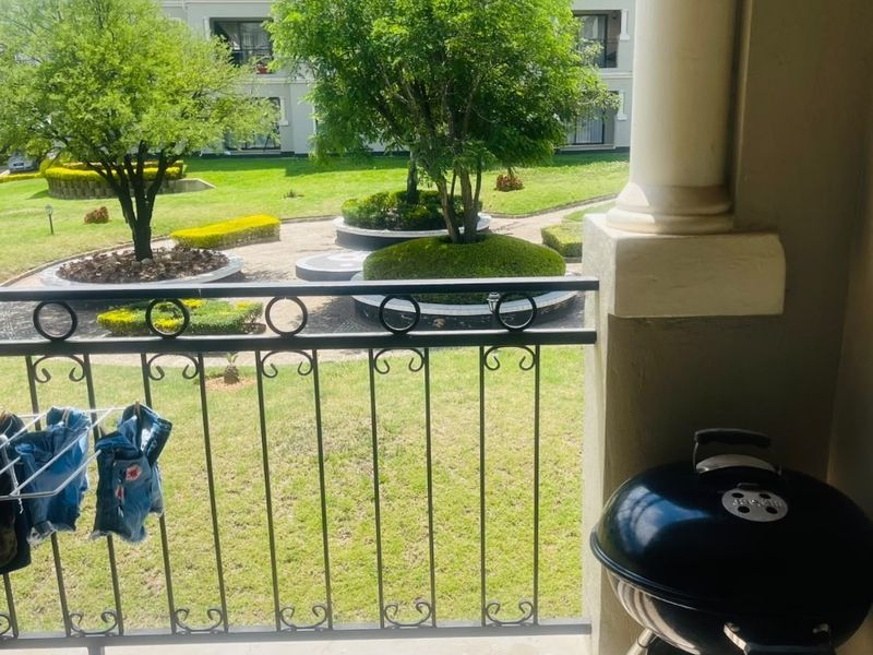 A covered balcony featuring tiled flooring and a wrought iron railing, overlooking a well-maintained communal garden area within the apartment complex.