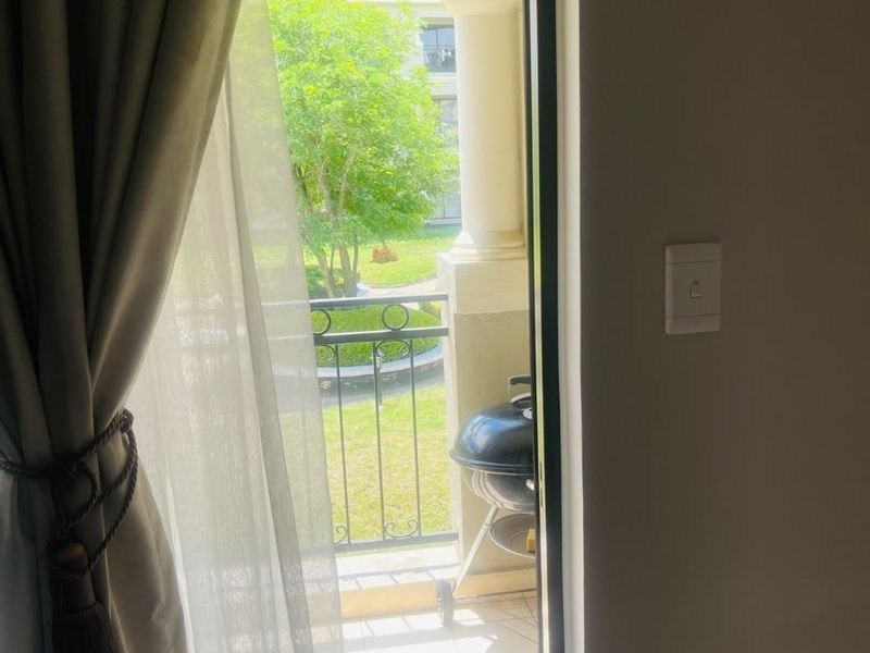 A view from an interior room looking out onto a private balcony featuring tiled flooring and a wrought iron railing overlooking a garden area.