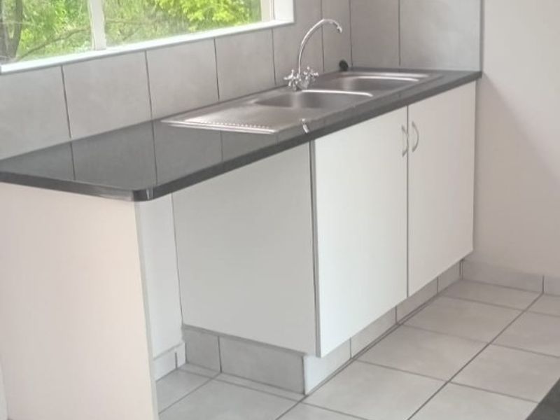A clean, functional kitchen area featuring a stainless steel sink, dark laminate countertops, white base cabinetry, and neutral ceramic floor tiles.