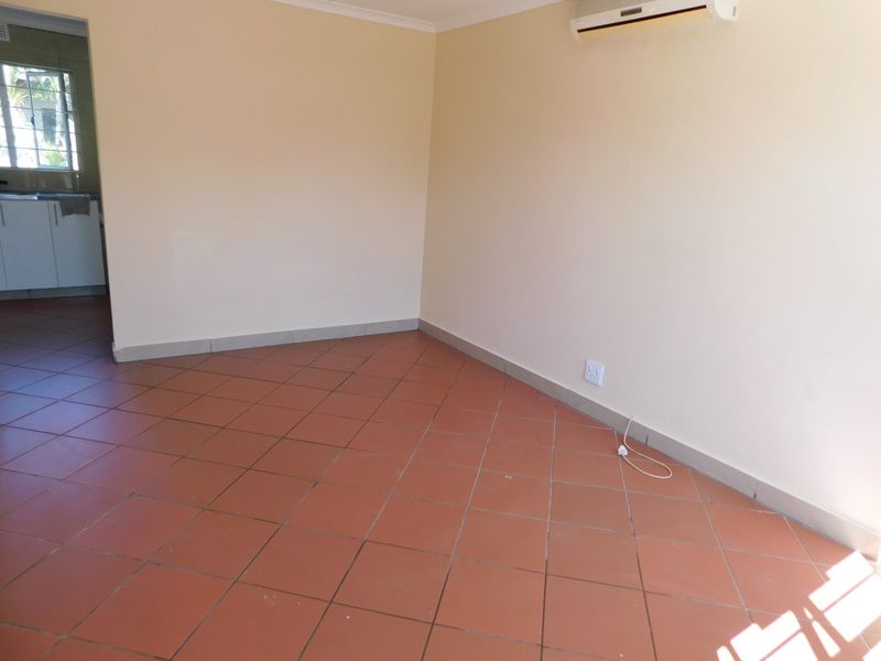 An empty living room featuring terracotta-style ceramic floor tiles, neutral wall paint, and a wall-mounted air conditioning unit, with a view into the adjacent kitchen.