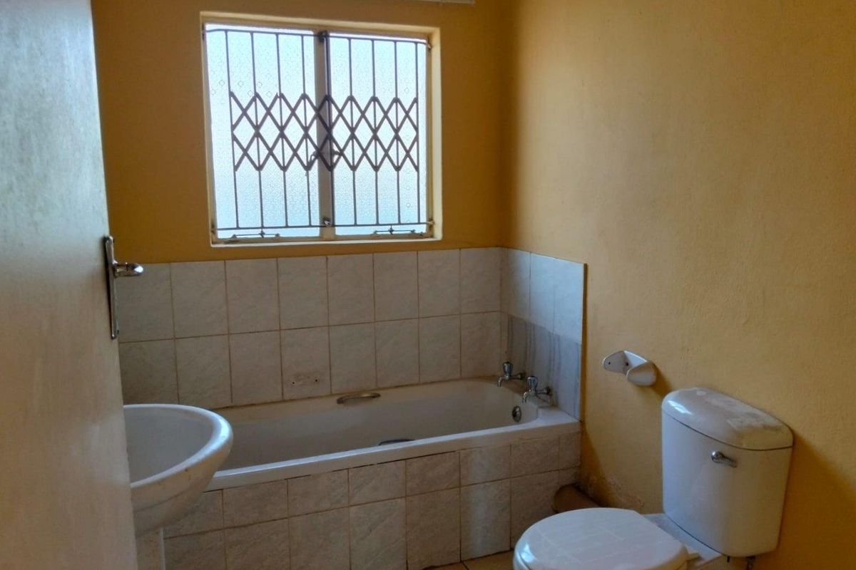 A functional bathroom featuring a bathtub, pedestal basin, and toilet, with security bars on the window and tiled finishes throughout.