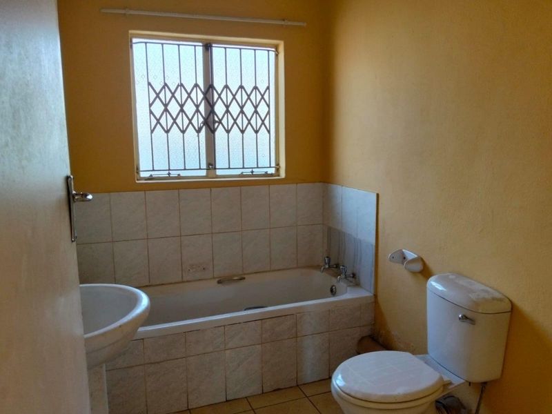 A functional bathroom featuring a bathtub, pedestal basin, and toilet, with security bars on the window and tiled finishes throughout.