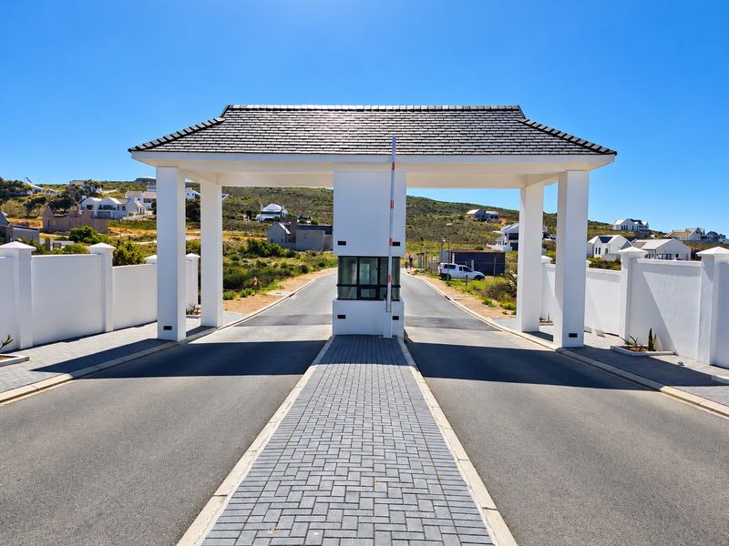 A well-maintained residential estate entrance featuring a modern security gatehouse, paved driveway, and perimeter walling under a clear blue sky.