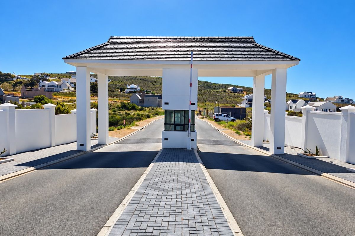 A well-maintained residential estate entrance featuring a modern security gatehouse, paved driveway, and perimeter walling under a clear blue sky.