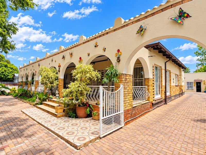 A charming front exterior featuring a covered arched veranda, decorative brickwork, and extensive paved driveway space with secure gated access.