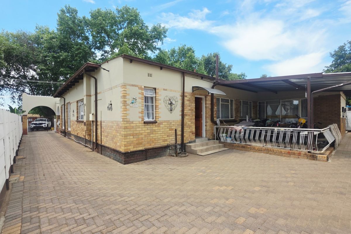 Rear exterior view of the property featuring a paved driveway, a covered patio area with metal railings, and a brick and plaster facade.