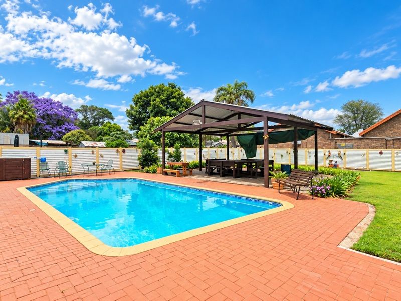 A well-maintained outdoor pool area featuring brick paving, a covered patio structure, and a spacious lawn, ideal for outdoor entertaining.