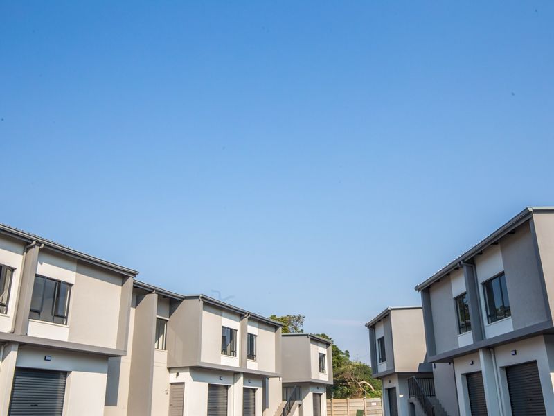 Modern apartment complex exterior featuring contemporary grey and white facade, roller shutter garage doors, and aluminium window frames under a clear blue sky.