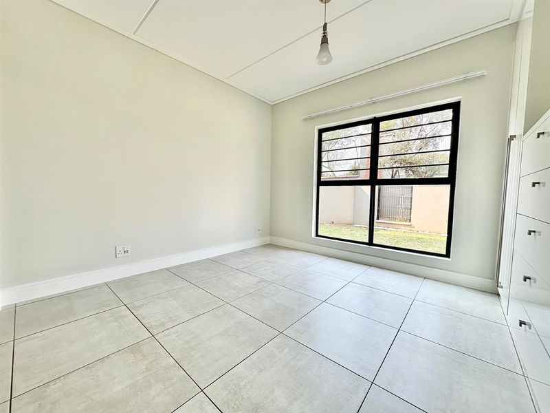 A bright, empty bedroom featuring modern large-format floor tiles, built-in cupboards, and a window with security bars overlooking the garden.