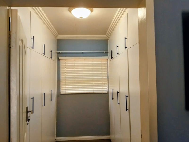 A clean, functional dressing area or bedroom walk-in closet featuring extensive built-in cabinetry, neutral wall tones, and a window with fitted venetian blinds.
