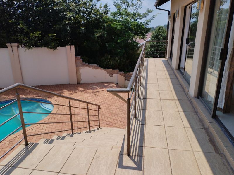 An elevated tiled balcony overlooking a paved patio area with a swimming pool, featuring metal railings and direct access via sliding glass doors.