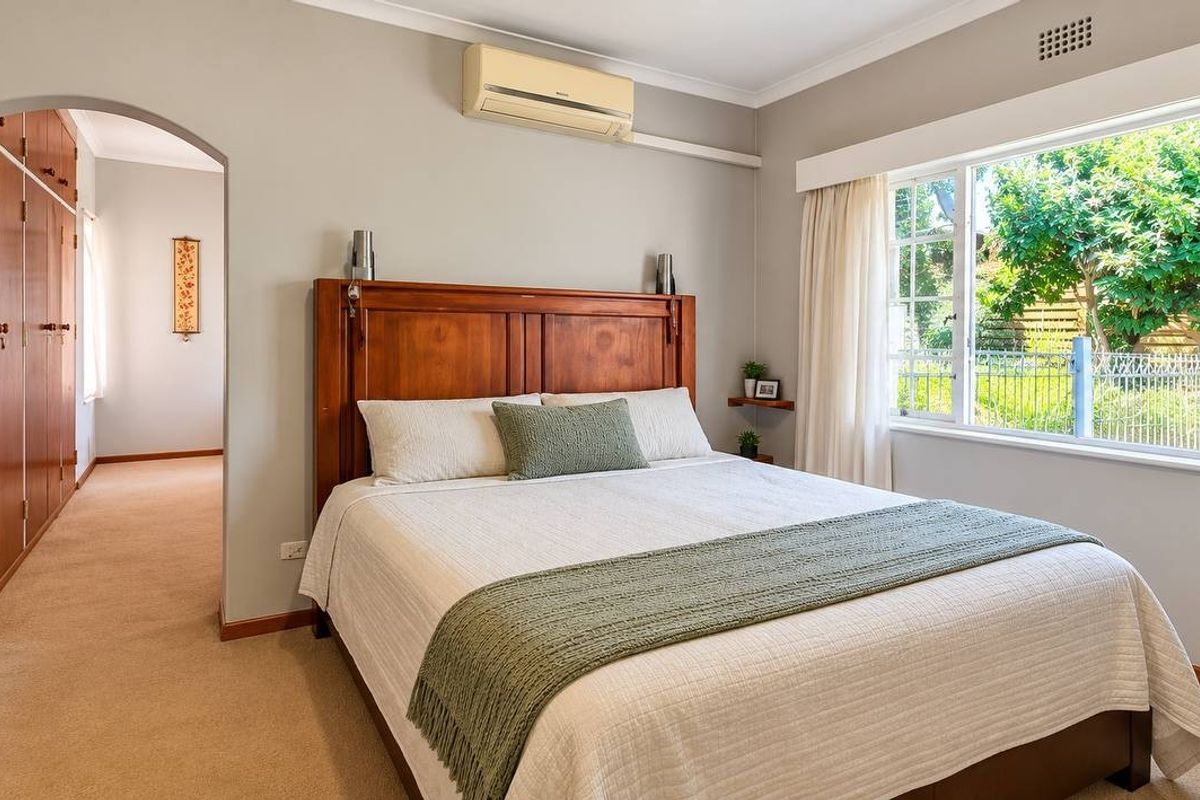 A well-maintained bedroom featuring built-in wooden cupboards, a wall-mounted air conditioning unit, and large windows providing natural light and garden views.