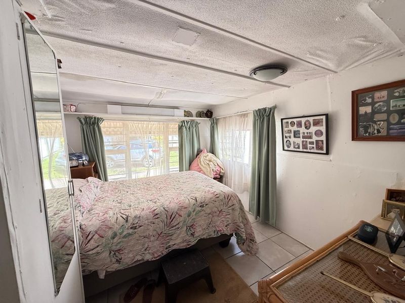 A bedroom featuring tiled flooring, a textured ceiling, and large windows allowing natural light. The space requires cosmetic updates to improve its overall condition.