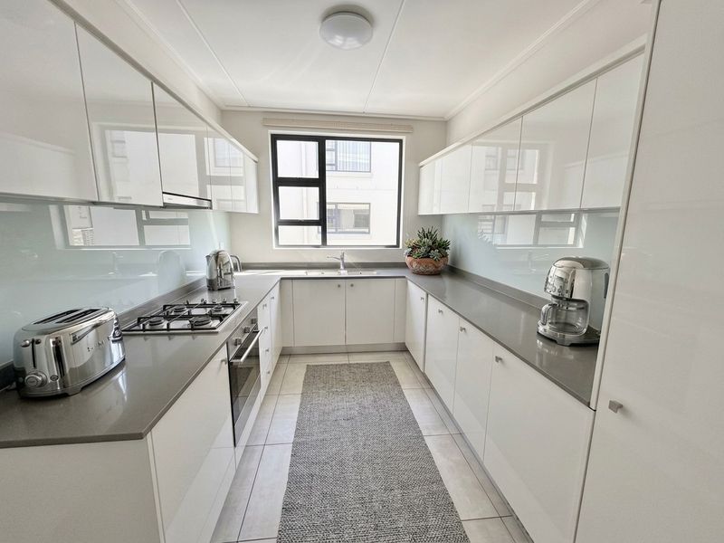 Modern galley-style kitchen featuring high-gloss white cabinetry, stone countertops, integrated gas hob, oven, and a sleek glass splashback.