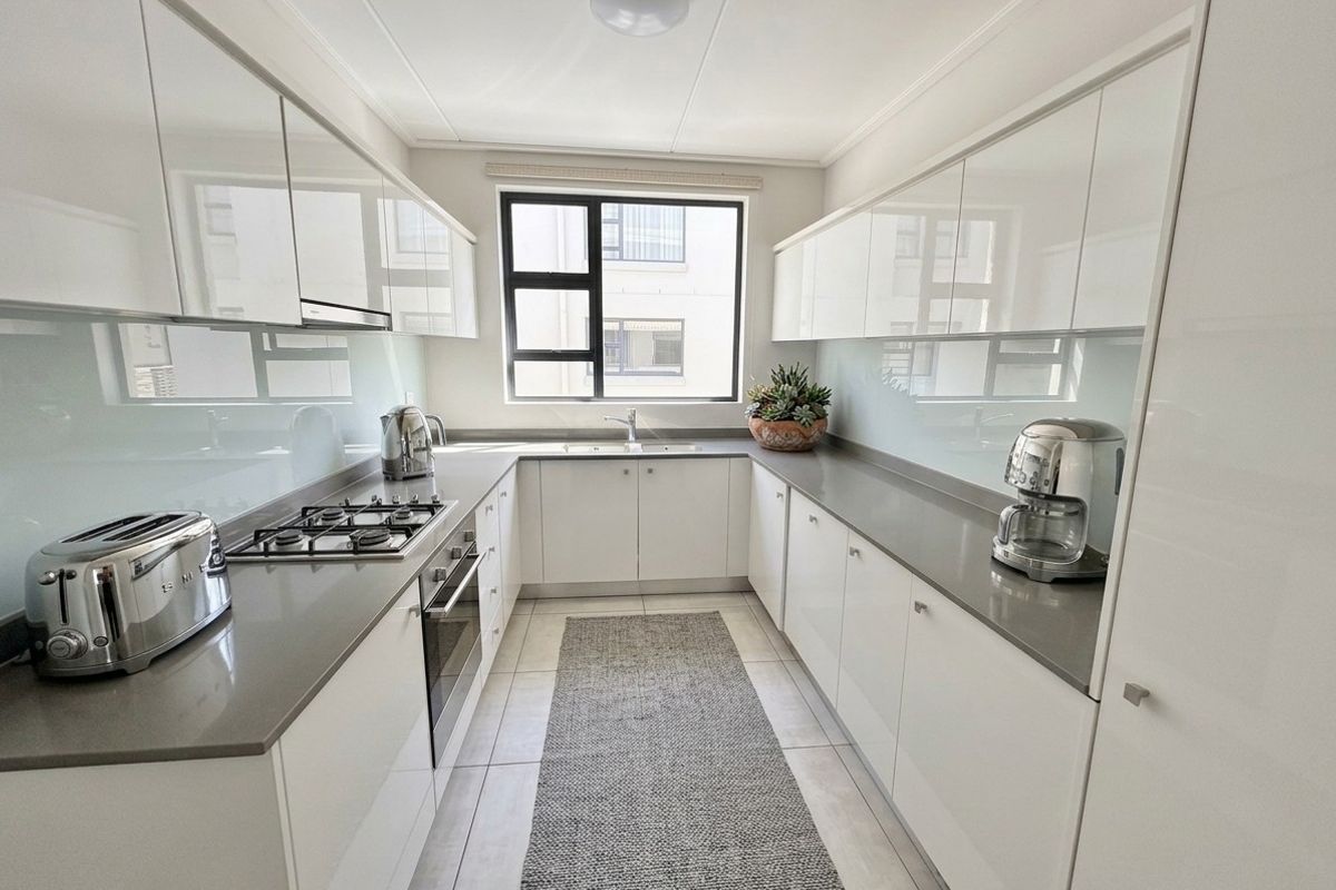 Modern galley-style kitchen featuring high-gloss white cabinetry, stone countertops, integrated gas hob, oven, and a sleek glass splashback.