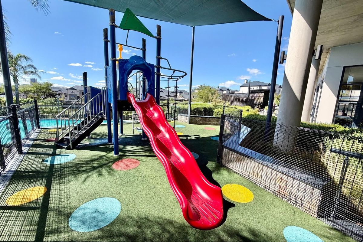 A well-maintained outdoor playground area featuring a slide, climbing structure, shade sail, and synthetic turf flooring within the apartment complex.