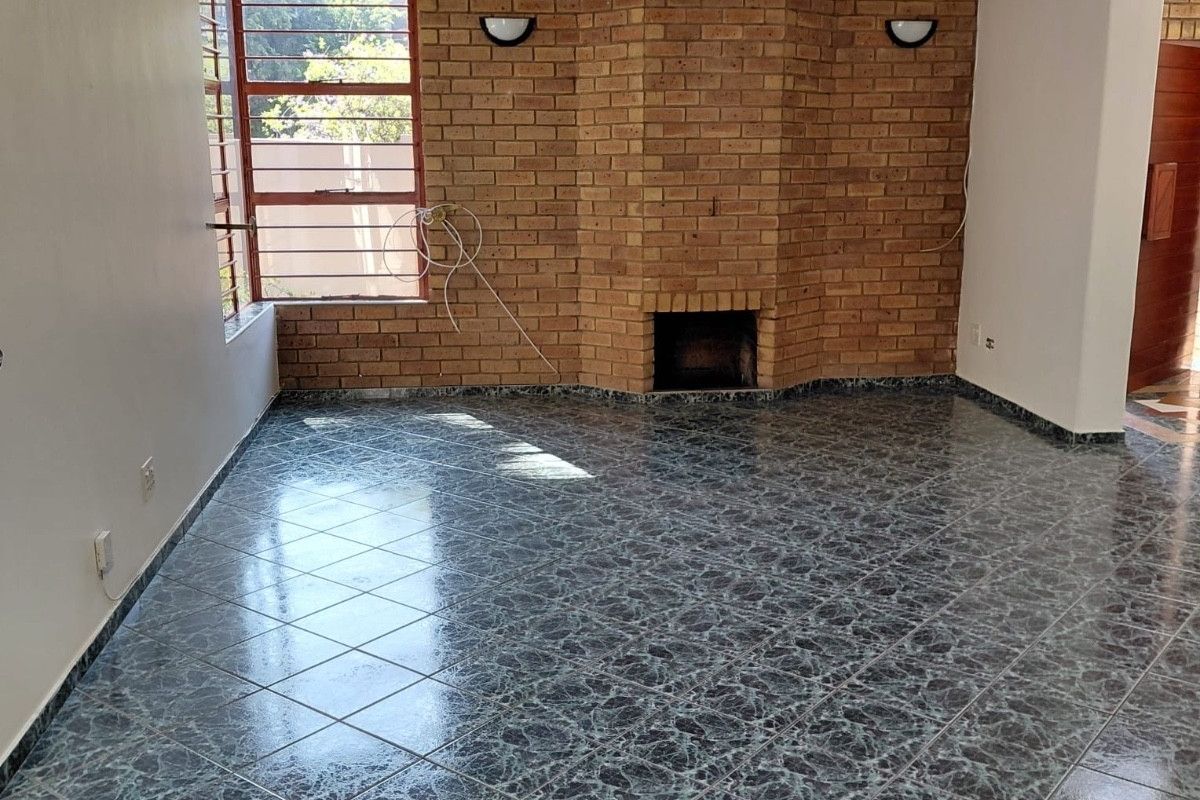 An empty living room featuring a prominent brick fireplace, tiled flooring, and large windows with steel frames, offering a blank canvas for renovation.