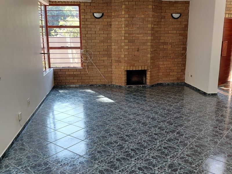 An empty living room featuring a prominent brick fireplace, tiled flooring, and large windows with steel frames, offering a blank canvas for renovation.