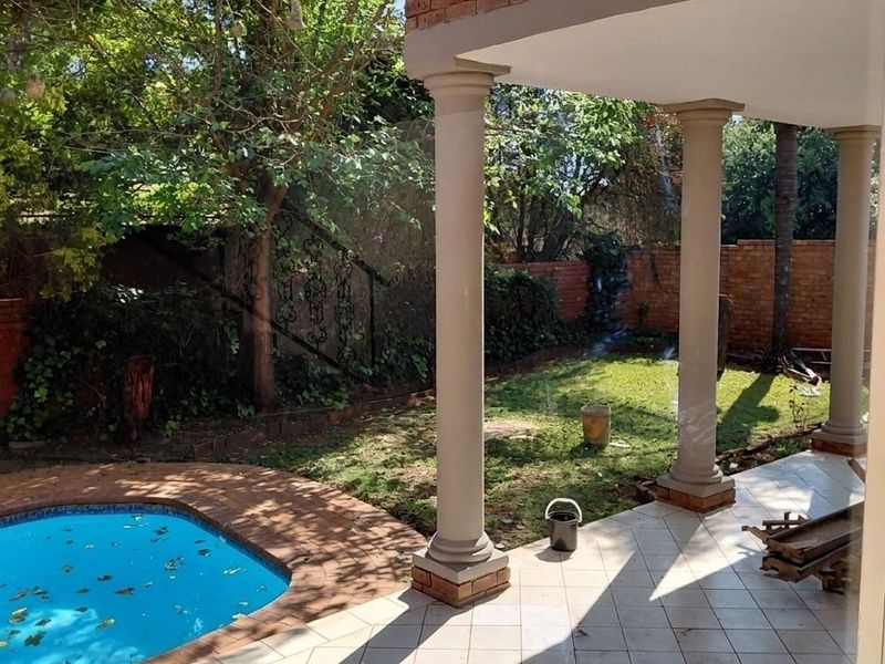 Outdoor patio area featuring tiled flooring and structural columns, overlooking a private swimming pool and garden space with brick boundary walls.