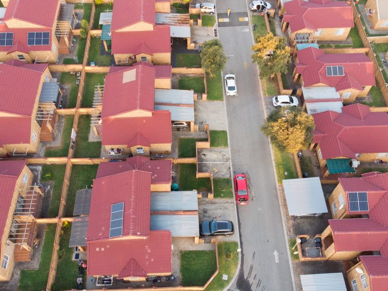 An aerial view of a residential estate featuring multiple detached houses with red tiled roofs, solar panels, and private garden plots.