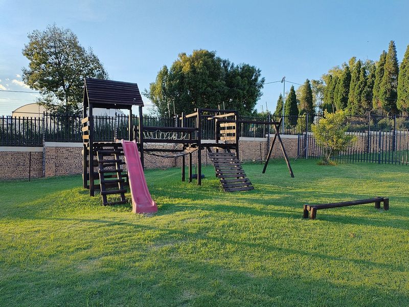 A well-maintained communal garden area featuring a wooden playground structure, manicured lawn, and secure perimeter fencing.