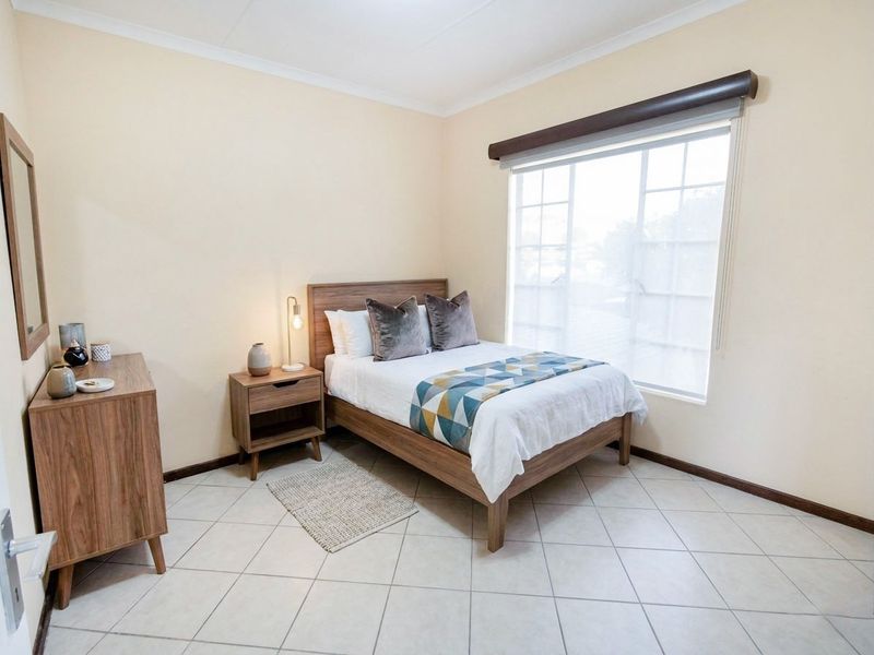 A well-maintained bedroom featuring neutral wall tones, tiled flooring, and a large window with a roller blind, providing ample natural light.