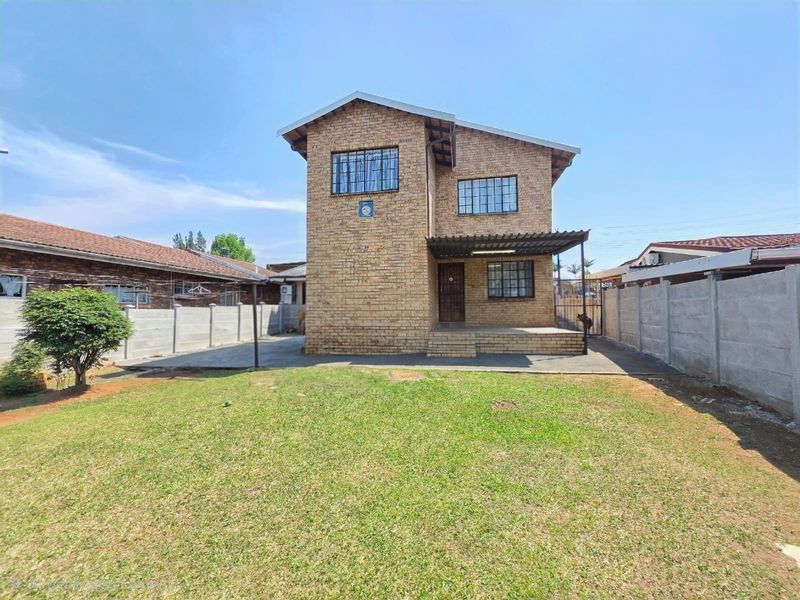 A rear view of a double-story face brick house featuring a concrete patio, security bars on windows, and a spacious private garden with boundary walls.
