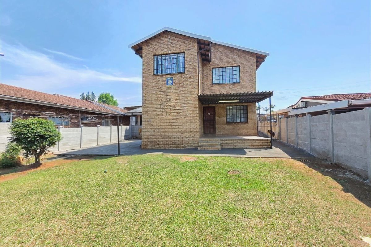 A rear view of a double-story face brick house featuring a concrete patio, security bars on windows, and a spacious private garden with boundary walls.
