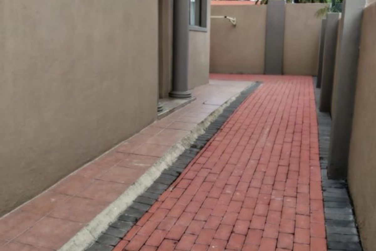 A side walkway featuring red brick paving, bordered by a plastered boundary wall and the side elevation of the house.