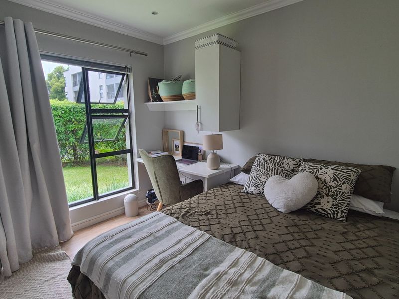 A well-lit bedroom featuring a built-in desk and shelving unit, with large windows overlooking a garden area.