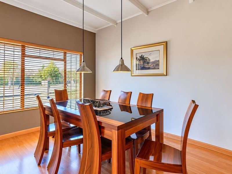 A well-lit dining room featuring laminate flooring, exposed ceiling beams, and large windows with timber frames and venetian blinds.