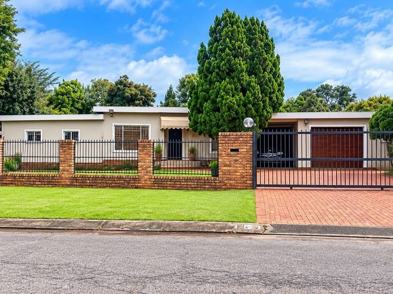 A well-maintained single-story house featuring a brick boundary wall, metal security fencing, a paved driveway, and a double garage with an automated gate.