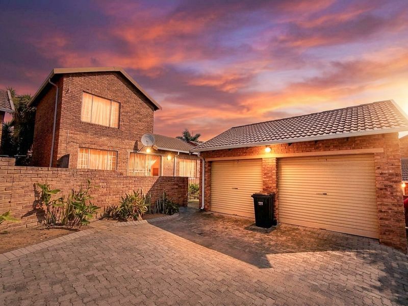 Front exterior view of a brick townhouse featuring a double garage, paved driveway, and boundary wall at sunset.