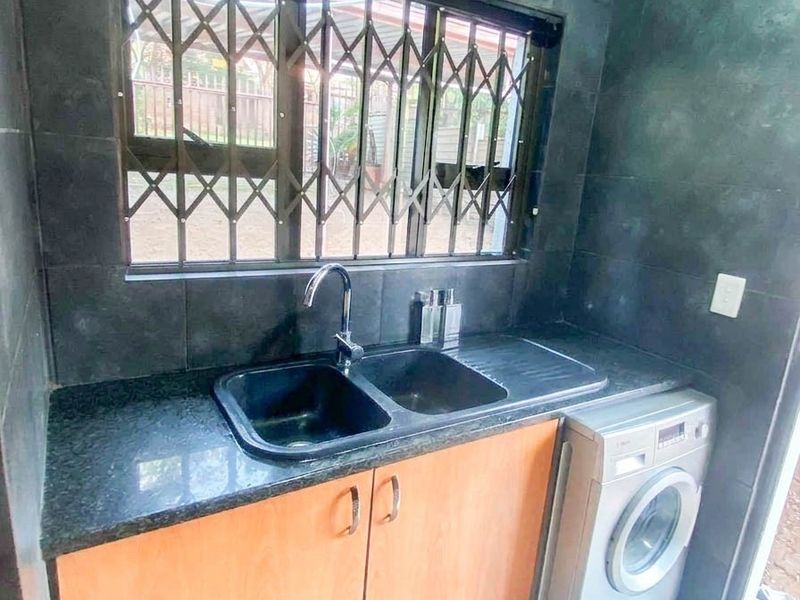 A functional laundry area featuring dark tiled walls, granite countertops, a double basin sink, and dedicated space for a washing machine.