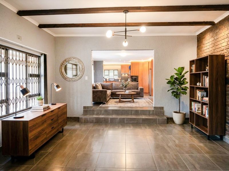 Spacious living area featuring exposed timber ceiling beams, tiled flooring, and a brick feature wall, with a raised transition into the adjacent kitchen area.