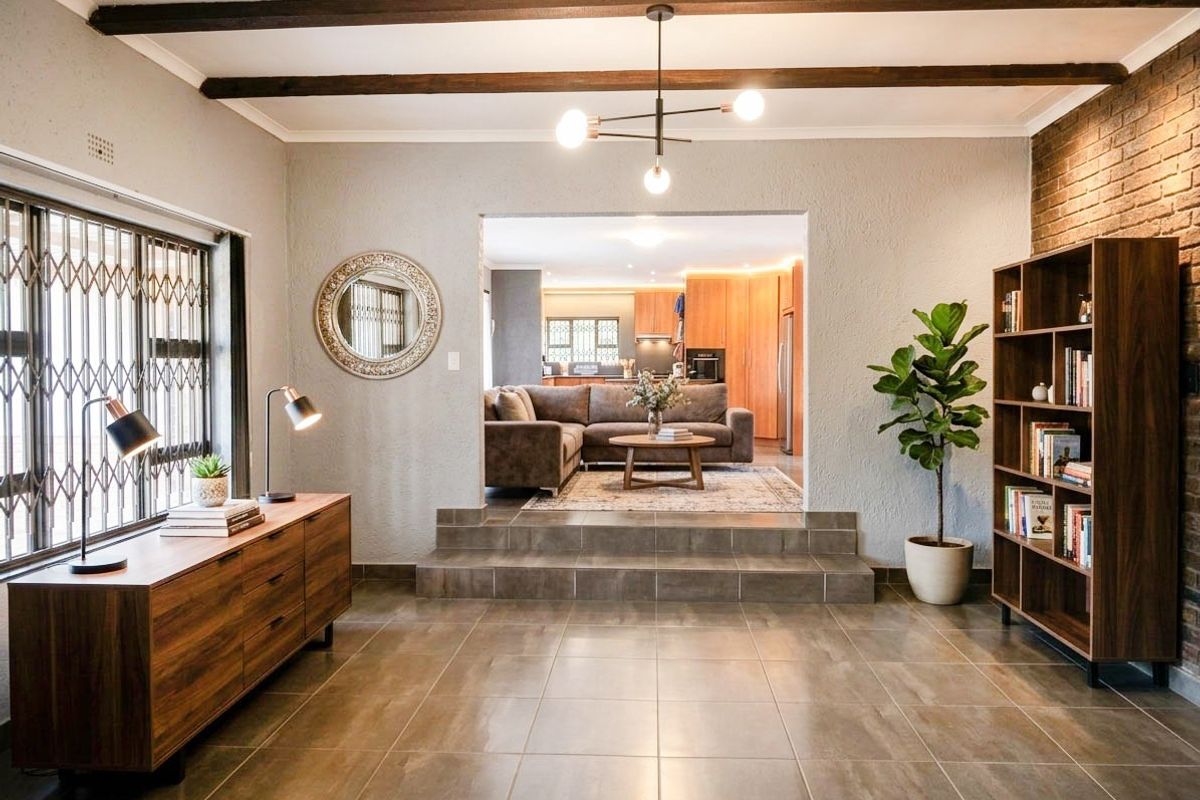 Spacious living area featuring exposed timber ceiling beams, tiled flooring, and a brick feature wall, with a raised transition into the adjacent kitchen area.