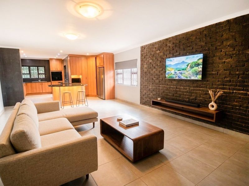 Spacious open-plan living area featuring a striking exposed brick wall, modern kitchen with wooden cabinetry, and sleek tiled flooring throughout.