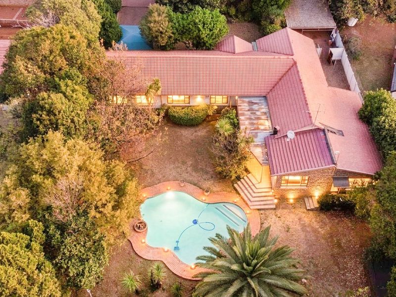 Aerial view of a residential property featuring a large L-shaped house with a tiled roof, a swimming pool, and established garden surroundings.