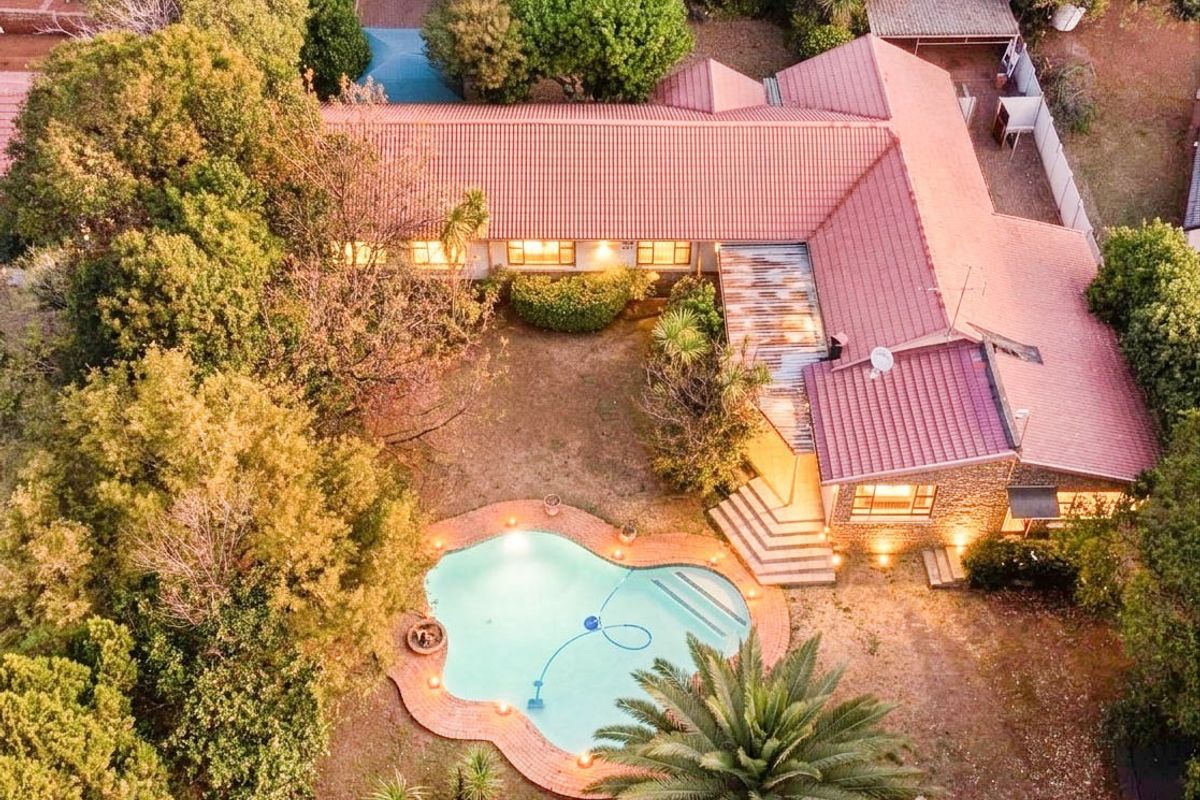 Aerial view of a residential property featuring a large L-shaped house with a tiled roof, a swimming pool, and established garden surroundings.