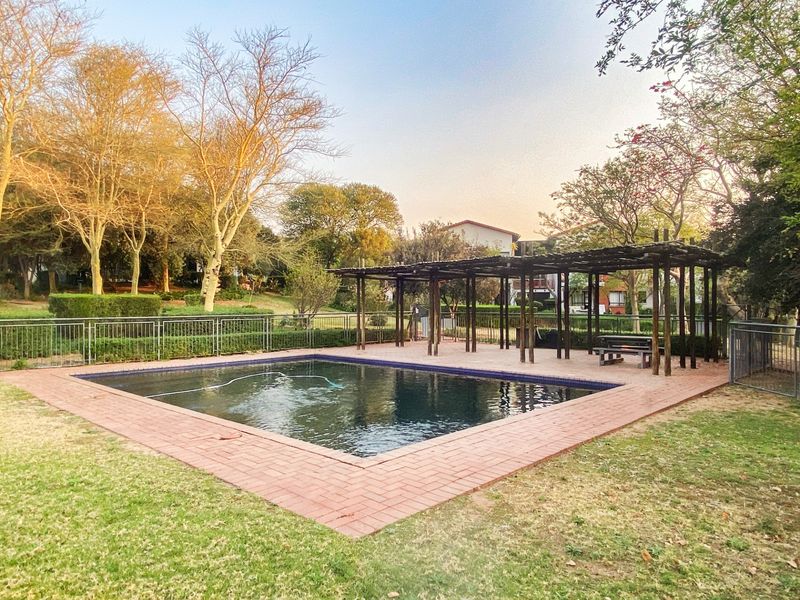 Communal swimming pool area featuring a brick-paved surround, a large timber pergola for shade, and secure metal perimeter fencing set within landscaped grounds.