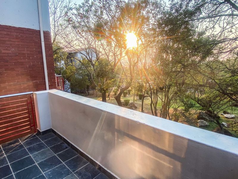 A private balcony featuring tiled flooring, a brick exterior wall, and a metal balustrade, overlooking a landscaped garden area.