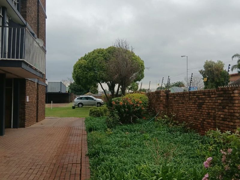 Rear exterior view of the apartment building featuring a paved walkway, brick boundary wall with electric fencing, and a garden area.
